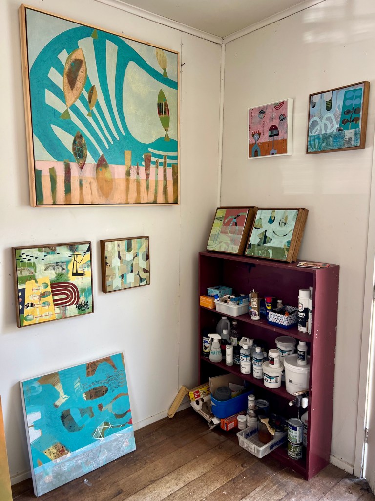 Photo of a corner in the artist's studio showing small works on the wall and a shelf filled with paint and other equipment